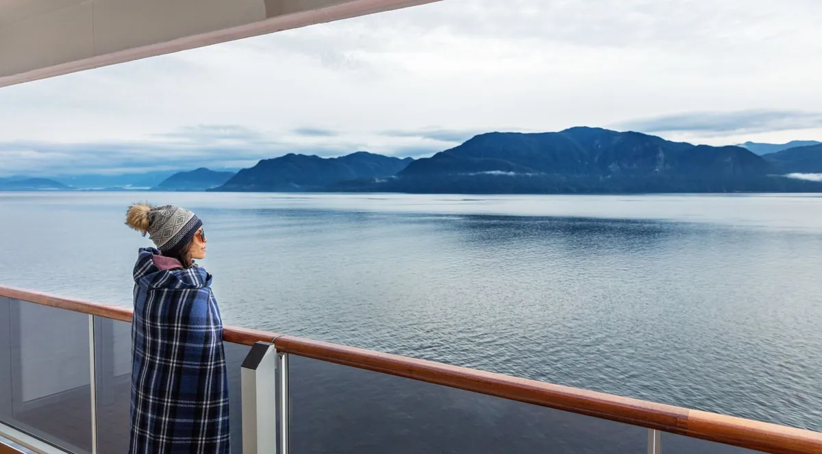 A traveler wrapped in a blanket on a boat deck watching a calm fjord