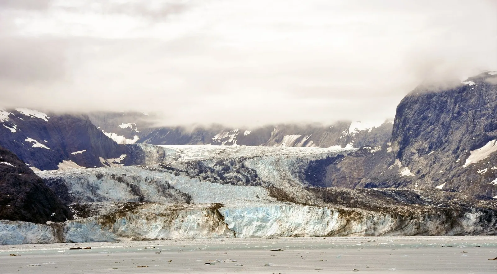 A glacier flowing down a valley between cloud-wrapped mountains